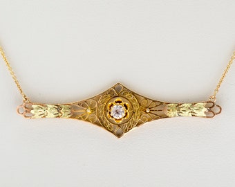 May include: A gold-toned necklace with a filigree design. The pendant features a central diamond and leaf accents. The necklace has a delicate chain and is set against a white background.