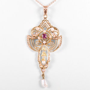 May include: A gold-toned pendant necklace with a central ruby and surrounding pearls. The pendant features an ornate, symmetrical design with delicate filigree and a dangling pearl drop. The chain is a fine, rose gold color.