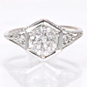 Könnte beinhalten: Ein Verlobungsring im Vintage-Stil mit einem großen, funkelnden Diamanten in einem sechseckigen Rahmen. Das Weißgoldband weist zarte florale und geometrische Gravuren auf. Das Design des Rings ist elegant und zeitlos und zeigt eine klassische Ästhetik.