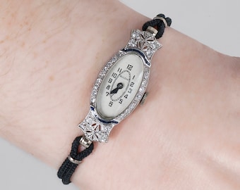 May include: A vintage Bulova wristwatch with a white face and black hands. The watch is set in a silver oval case with diamond accents and a black fabric band.