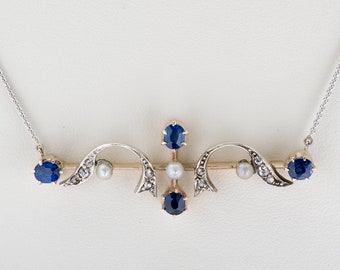 May include: A silver necklace with a pendant featuring blue sapphires, small pearls, and clear gemstones. The pendant has a symmetrical design with a central bar and curved accents. The necklace is set against a white background.