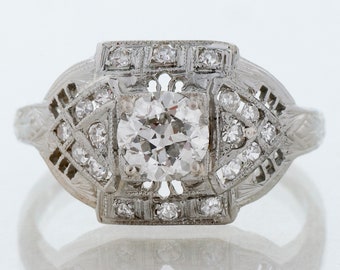 May include: A vintage platinum engagement ring with a round brilliant cut diamond center stone and a geometric design with smaller diamonds surrounding the center stone.