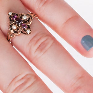 Puede incluir: Anillo dorado con dise&ntilde;o floral, engastado con piedras preciosas de color rojo oscuro y peque&ntilde;as perlas blancas. Los intrincados detalles del anillo se destacan sobre un fondo claro. Estilo vintage.