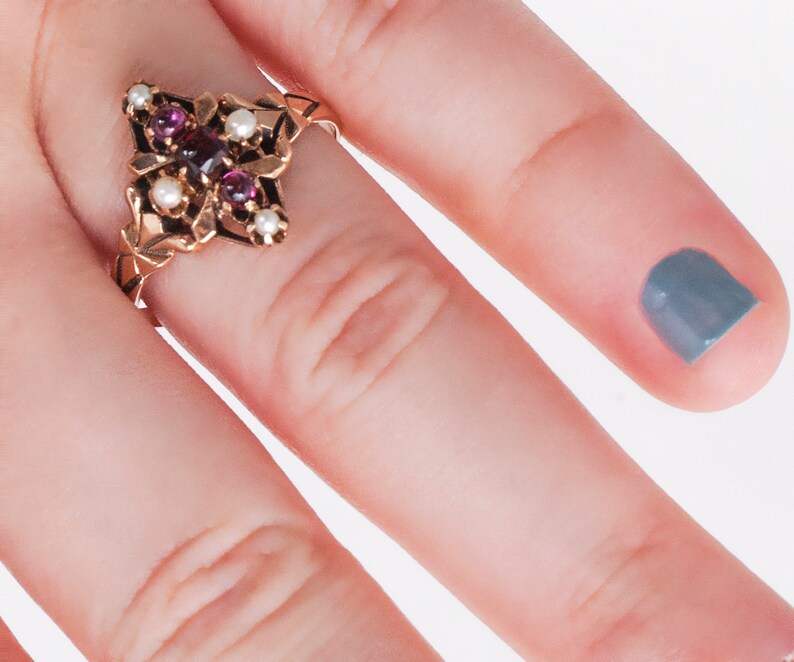 Puede incluir: Anillo dorado con dise&ntilde;o floral, engastado con piedras preciosas de color rojo oscuro y peque&ntilde;as perlas blancas. Los intrincados detalles del anillo se destacan sobre un fondo claro. Estilo vintage.