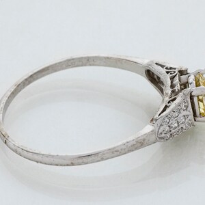 May include: A silver ring with a large, yellow gemstone and smaller clear stones. The ring has an ornate design on the sides of the gemstone and a simple band. The ring is set against a white background.