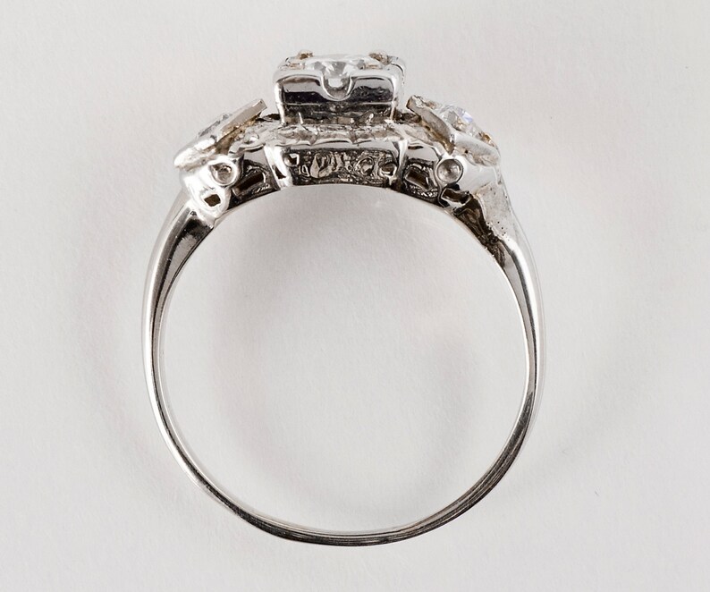 Puede incluir: Un anillo de plata con una piedra central grande de forma cuadrada y piedras m&aacute;s peque&ntilde;as a cada lado. El anillo est&aacute; engastado en un dise&ntilde;o delicado e intrincado.