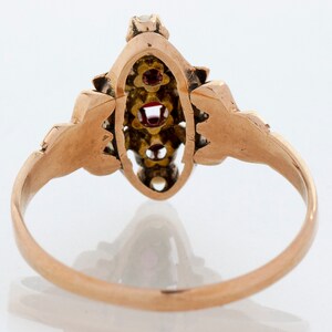 Puede incluir: Un anillo de oro con una pieza central de forma ovalada. El anillo presenta tres piedras preciosas rojas y varias piedras preciosas claras m&aacute;s peque&ntilde;as. La banda y los elementos decorativos tienen un dise&ntilde;o detallado y ornamentado.