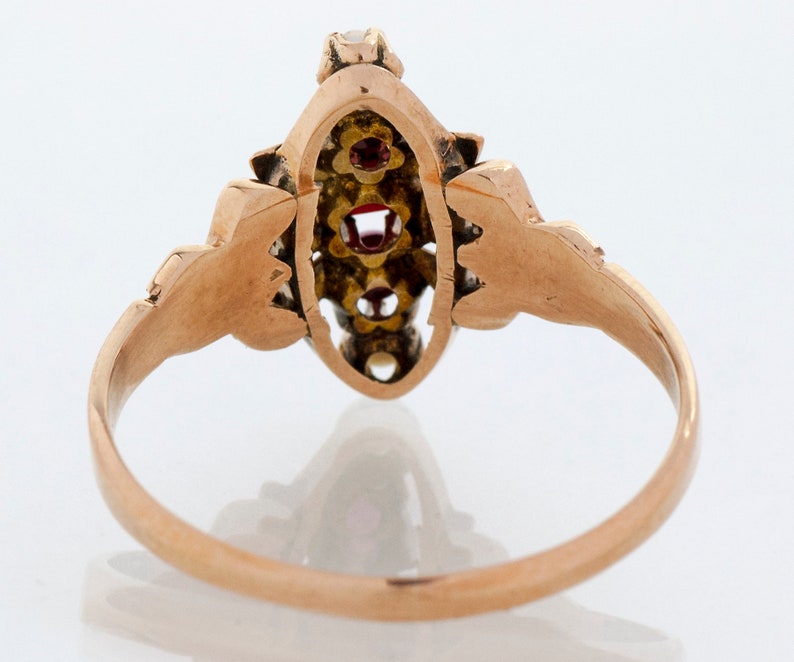 Puede incluir: Un anillo de oro con una pieza central de forma ovalada. El anillo presenta tres piedras preciosas rojas y varias piedras preciosas claras m&aacute;s peque&ntilde;as. La banda y los elementos decorativos tienen un dise&ntilde;o detallado y ornamentado.