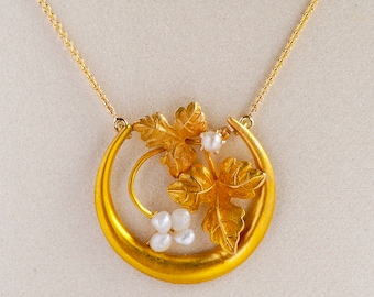 May include: A gold-toned necklace featuring a circular pendant with a stylized leaf and pearl design. The pendant has a crescent shape with a detailed leaf motif and small, white pearls. The necklace chain is also gold-toned.