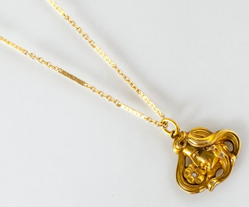 May include: A gold necklace with a pendant featuring a detailed, stylized image of a woman's face with a flower.