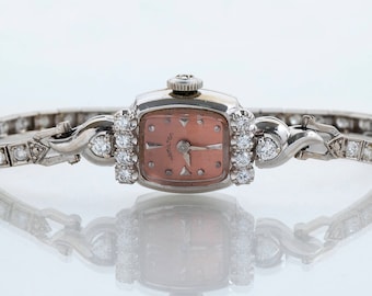 May include: A vintage wristwatch with a square face and a diamond-encrusted band. The watch face is a pale pink color with black hands and Roman numeral markings. The watch is set in a silver-toned metal.
