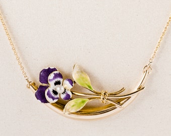 May include: A gold necklace with a purple and white pansy flower pendant. The flower is set on a gold stem with green leaves. The necklace is on a white background.
