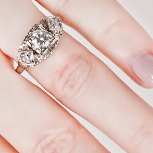 Puede incluir: Un anillo de plata con un gran diamante de talla cuadrada en el centro y dos diamantes m&aacute;s peque&ntilde;os a cada lado. El anillo est&aacute; engastado en un dise&ntilde;o delicado e intrincado.