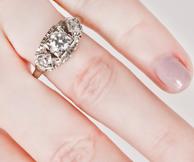 Puede incluir: Un anillo de plata con un gran diamante de talla cuadrada en el centro y dos diamantes m&aacute;s peque&ntilde;os a cada lado. El anillo est&aacute; engastado en un dise&ntilde;o delicado e intrincado.