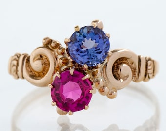 Puede incluir: Un anillo de oro con dos piedras preciosas, una azul oscuro y otra magenta. El anillo tiene un diseño de banda ornamental y arremolinado. Una joya llamativa con un diseño clásico.