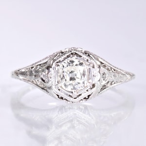 Könnte beinhalten: Ein Vintage-Verlobungsring mit einem zentralen runden Brillanten, der von kleineren Diamanten umgeben ist. Der Ring ist in Platin gefasst und hat ein filigranes Design.