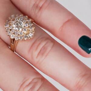 May include: A gold ring featuring a large, round, clear center stone surrounded by smaller, clear stones in a floral or starburst pattern. The ring is on a finger with a dark teal nail.