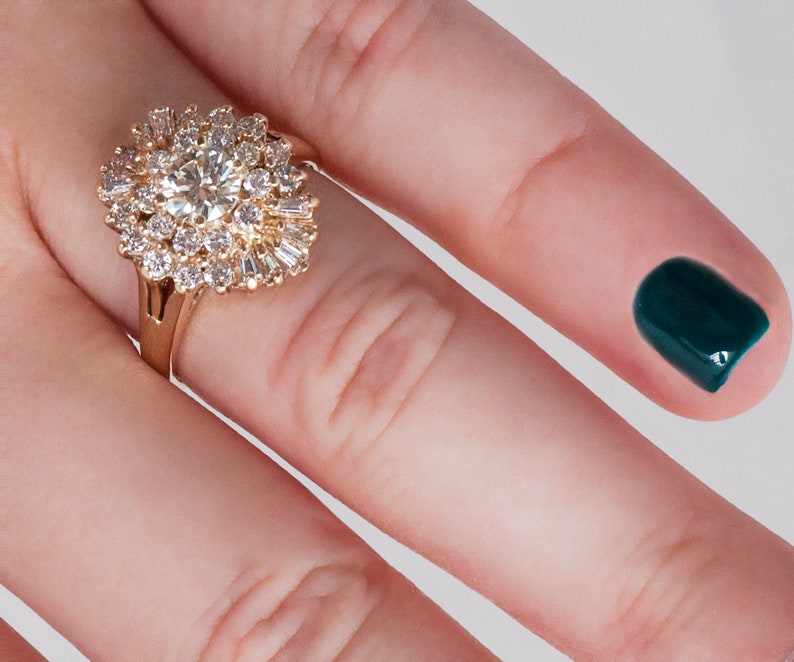 May include: A gold ring featuring a large, round, clear center stone surrounded by smaller, clear stones in a floral or starburst pattern. The ring is on a finger with a dark teal nail.