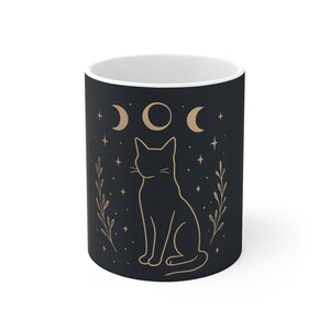 May include: A black ceramic mug featuring a light brown line art illustration of a seated cat centered on the front. Above the cat are three moon phases, flanked by vertical leafy branches and small stars scattered across the dark surface.