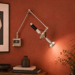 May include: A wall-mounted articulated lamp with a polished chrome finish, featuring a square base, an adjustable metal arm, and a cylindrical black and chrome light head. The lamp is positioned to cast light downward, highlighting its mechanical joints.