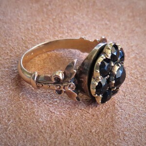 May include: A gold ring with a black stone cluster set in a bezel setting. The ring has a decorative band with a fleur-de-lis design.