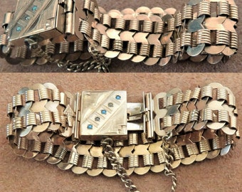 Puede incluir: Un brazalete de oro con un cierre decorativo con piedras azules. El brazalete está hecho de múltiples filas de piezas de metal enlazadas.