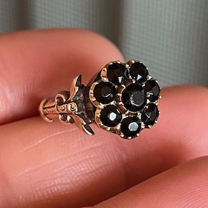 May include: A gold ring featuring a black flower-shaped cluster of gemstones. The ring has an intricate design on the band.