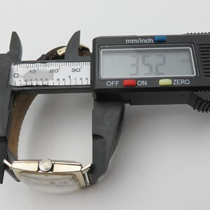 May include: A vintage wristwatch with a gold-colored rectangular case and a brown leather strap is measured with a digital caliper. The caliper displays a measurement of 35.2 mm, approximately 1.39 inches. The watch face is visible.