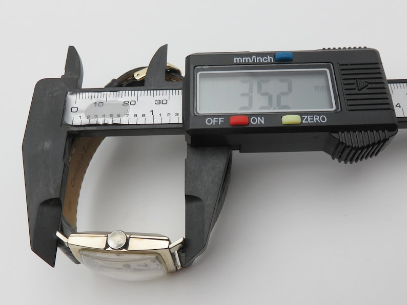 May include: A vintage wristwatch with a gold-colored rectangular case and a brown leather strap is measured with a digital caliper. The caliper displays a measurement of 35.2 mm, approximately 1.39 inches. The watch face is visible.