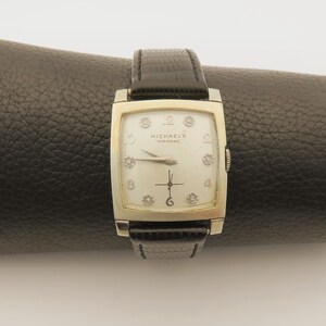 May include: A vintage rectangular watch with a silver-colored case and a black leather band. The watch face is off-white with diamond hour markers and the text "MICHAEL'S DIAMOND". The watch is displayed on a black surface.