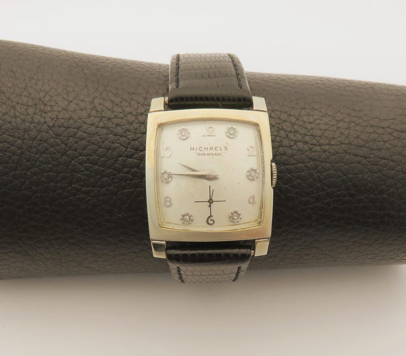 May include: A vintage rectangular watch with a silver-colored case and a black leather band. The watch face is off-white with diamond hour markers and the text "MICHAEL'S DIAMOND". The watch is displayed on a black surface.