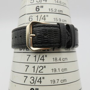 May include: Close-up of a black watch band with a gold-colored buckle. The band has a textured, reptile-like pattern. The watch band is measured on a white cone with inch and centimeter markings.