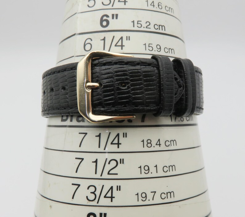 May include: Close-up of a black watch band with a gold-colored buckle. The band has a textured, reptile-like pattern. The watch band is measured on a white cone with inch and centimeter markings.
