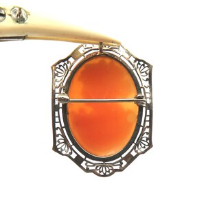 May include: A vintage cameo brooch with an oval, orange-colored center set in a filigree metal frame. The brooch has a pin closure on the back and is held by a tool.