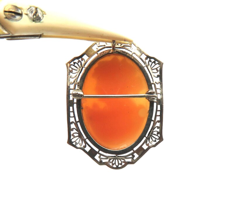 May include: A vintage cameo brooch with an oval, orange-colored center set in a filigree metal frame. The brooch has a pin closure on the back and is held by a tool.