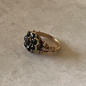 May include: A gold ring featuring a cluster of black gemstones set in the shape of a flower. The ring has a delicate design with intricate details.