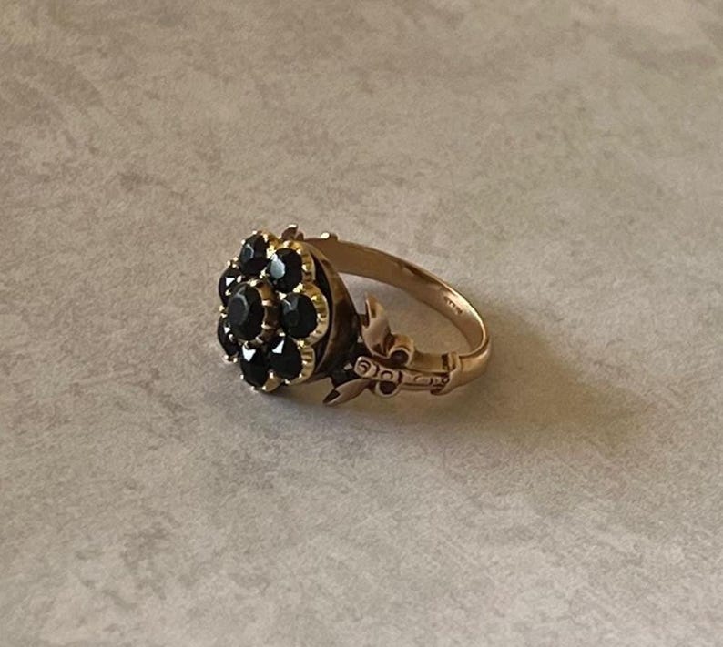 May include: A gold ring featuring a cluster of black gemstones set in the shape of a flower. The ring has a delicate design with intricate details.