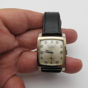 May include: A gold-toned rectangular watch with a black leather band. The watch face is cream-colored with diamond hour markers and the text "MICHAELS DIAMOND". The watch has a second hand and a crown on the right side.