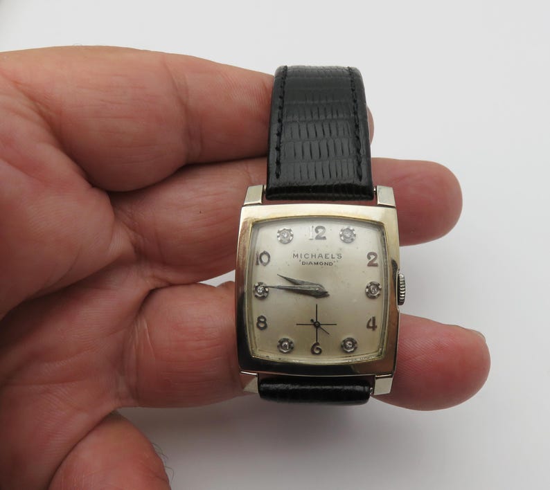 May include: A gold-toned rectangular watch with a black leather band. The watch face is cream-colored with diamond hour markers and the text "MICHAELS DIAMOND". The watch has a second hand and a crown on the right side.