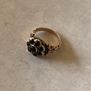 May include: A gold ring featuring a cluster of black gemstones in the centre. The ring has a floral design with intricate details.