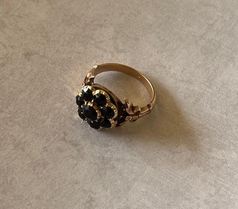 May include: A gold ring featuring a cluster of black gemstones in the centre. The ring has a floral design with intricate details.
