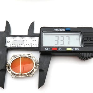 May include: An oval-shaped brooch with an orange center and a silver-colored filigree border. The brooch is measured with a digital caliper, showing a width of 33.7 mm. The caliper is black with a digital display.