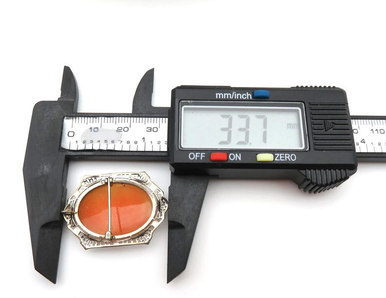 May include: An oval-shaped brooch with an orange center and a silver-colored filigree border. The brooch is measured with a digital caliper, showing a width of 33.7 mm. The caliper is black with a digital display.