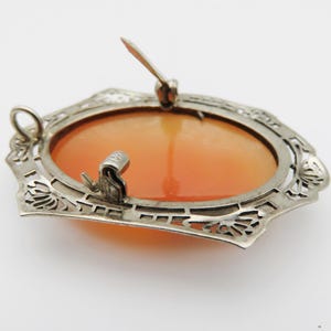 May include: A vintage cameo brooch with an oval, orange-toned center. The brooch is set in a silver-colored filigree frame with a pin closure on the back. The frame has a decorative, openwork design.
