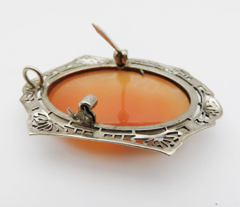 May include: A vintage cameo brooch with an oval, orange-toned center. The brooch is set in a silver-colored filigree frame with a pin closure on the back. The frame has a decorative, openwork design.