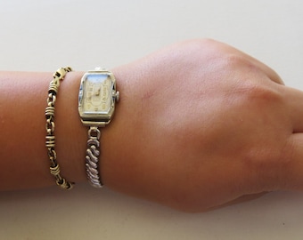 May include: A gold chain bracelet and a silver watch with a square face and a white dial on a person's wrist.