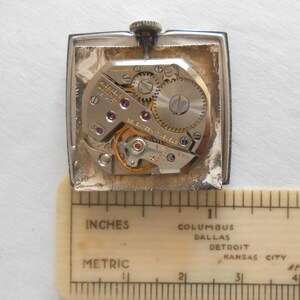 May include: A vintage wristwatch movement with visible gears, jewels, and intricate mechanical components. The watch is rectangular with a silver-colored case. A ruler with inch and metric measurements is placed below the watch.
