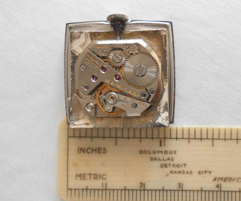 May include: A vintage wristwatch movement with visible gears, jewels, and intricate mechanical components. The watch is rectangular with a silver-colored case. A ruler with inch and metric measurements is placed below the watch.