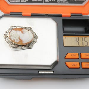 May include: An antique cameo brooch with a woman's profile carved in a light orange shell, set in a silver filigree frame. The brooch is on a digital scale, weighing 4.15 grams. The scale is black and orange.
