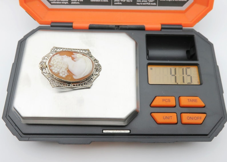 May include: An antique cameo brooch with a woman's profile carved in a light orange shell, set in a silver filigree frame. The brooch is on a digital scale, weighing 4.15 grams. The scale is black and orange.
